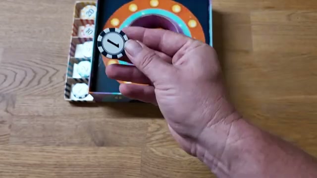 Hand hält einen Bounce-It-Punktechip mit der Zahl 1 über der Spielbox